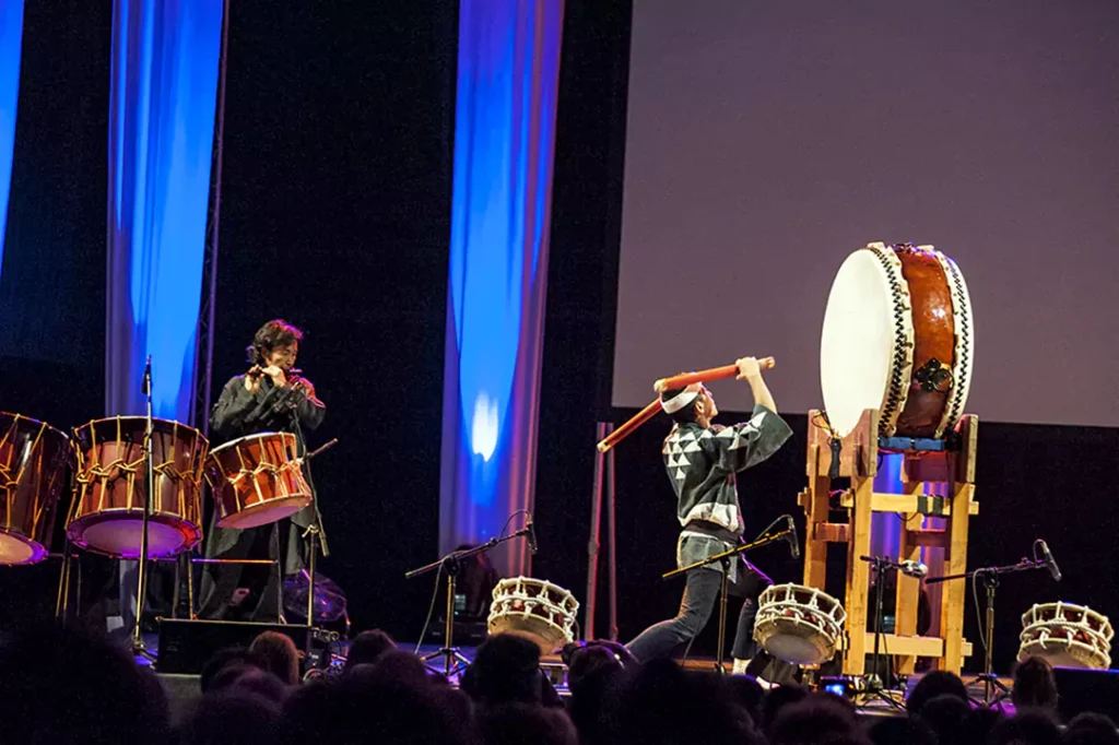 A close-up of an expressive taiko performer in colorful traditional attire, shouting while holding a drumstick forward in a powerful pose.