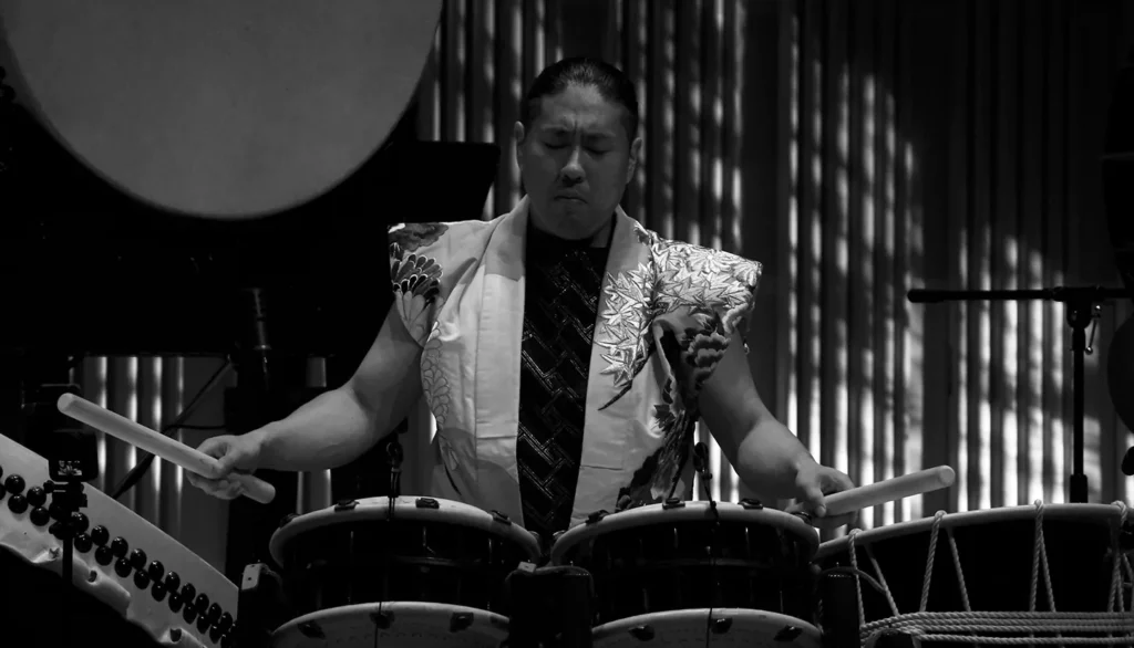 A black and white photo of a focused taiko drummer in traditional attire holding bachi over a set of drums, illustrating a guide on how to choose your taiko bachi.