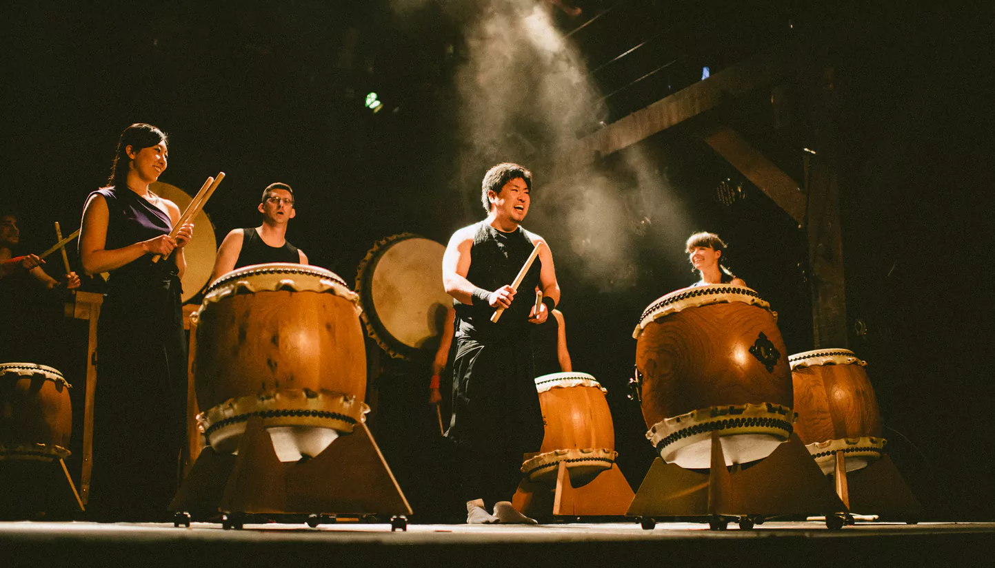 A diverse group of energetic performers playing large traditional wooden taiko drums on a dark, professional stage with theatrical lighting and smoke. An aspirational image for starting a school taiko team.