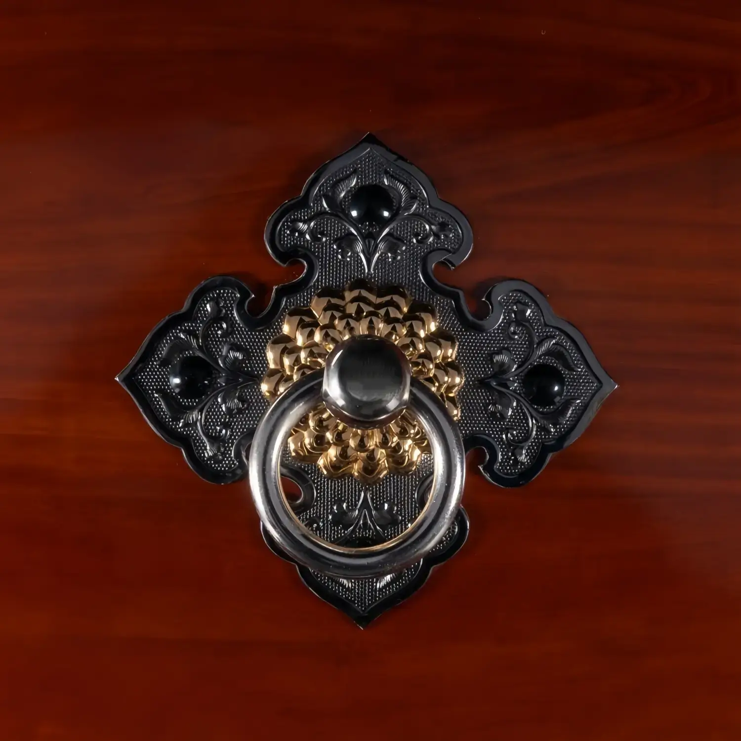 Close-up of JINLEI O-Daiko Akatsuki Series decorative hardware featuring gold and silver ring pull on ornate black cast iron diamond-shaped mount against deep red wood grain surface