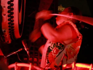 A live stage performance featuring a taiko drummer striking a large drum alongside a flute player, set against a blue backdrop with an audience watching.