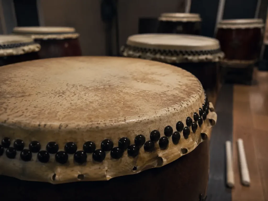 Close-up view of the worn leather texture on a Nagado Daiko drumhead and traditional black mounting tacks, with bachi resting in the blurred background.