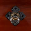 Close-up of O-Daiko Kapiac Do Series ornate hardware featuring gold floral ring pull on detailed black cast iron diamond-shaped mount against deep red kapiac wood grain surface