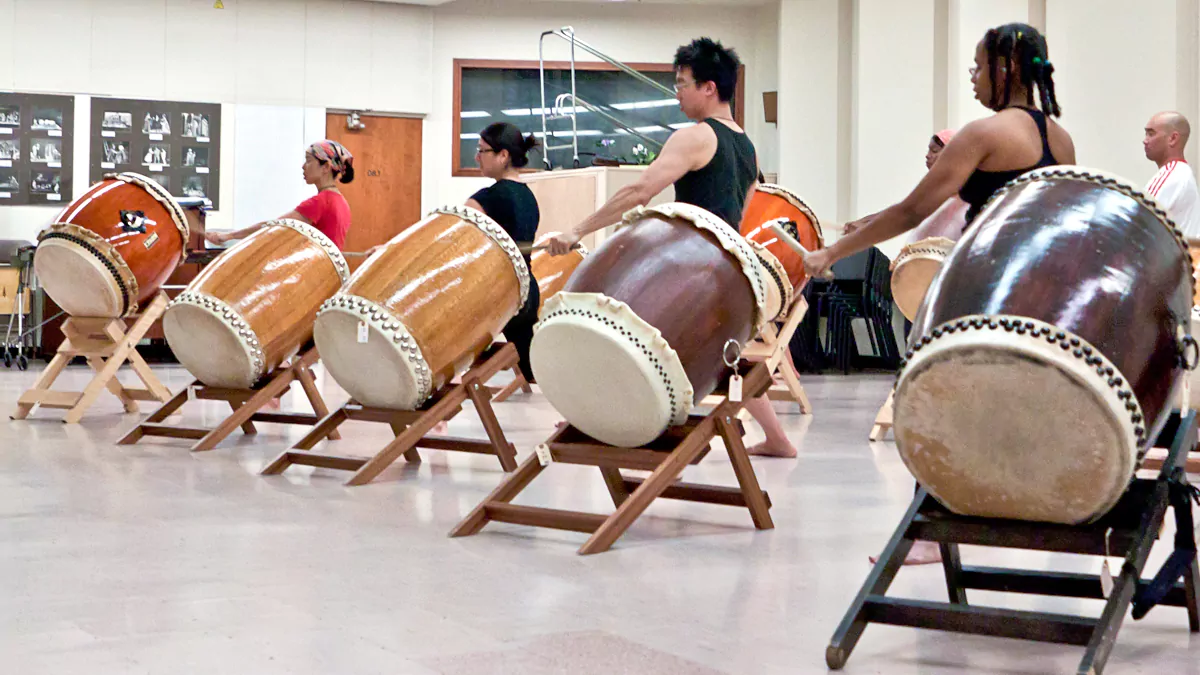 A group of students practicing taiko drumming on slant stands in a bright studio, illustrating systematic taiko teaching methods and group coordination.