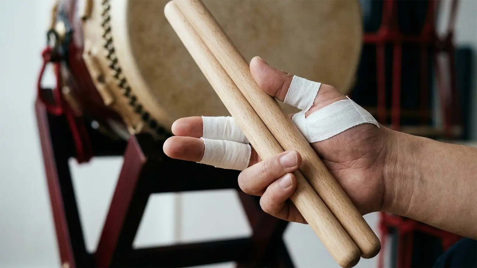 A cinematic close-up photo of a taiko player's hand with white breathable athletic tape applied to key fingers, demonstrating a relaxed grip on a pair of natural wood bachi. In the softly blurred background are a taiko drum and its dark red stand.