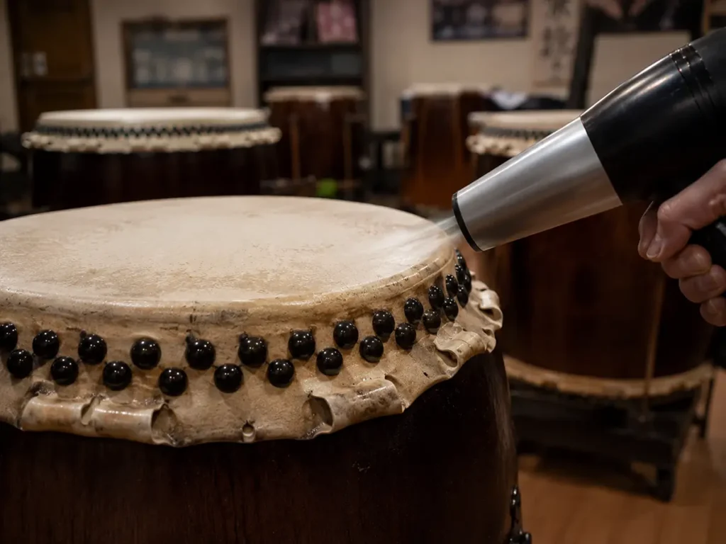 A person using a heat gun to warm the leather surface of a Nagado Daiko drum, a common maintenance technique for adjusting tension and tuning.