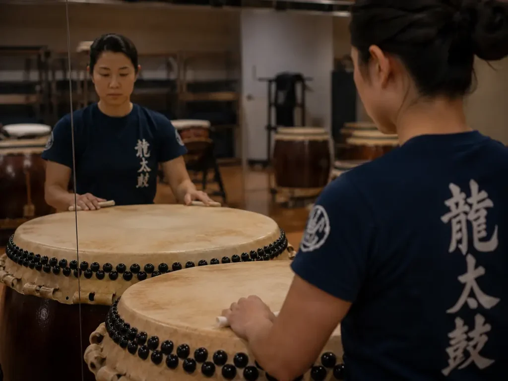 A female taiko drummer sitting in front of a mirror during practice, with her hands resting on the Nagado Daiko drumhead and her focused expression reflected in the glass.