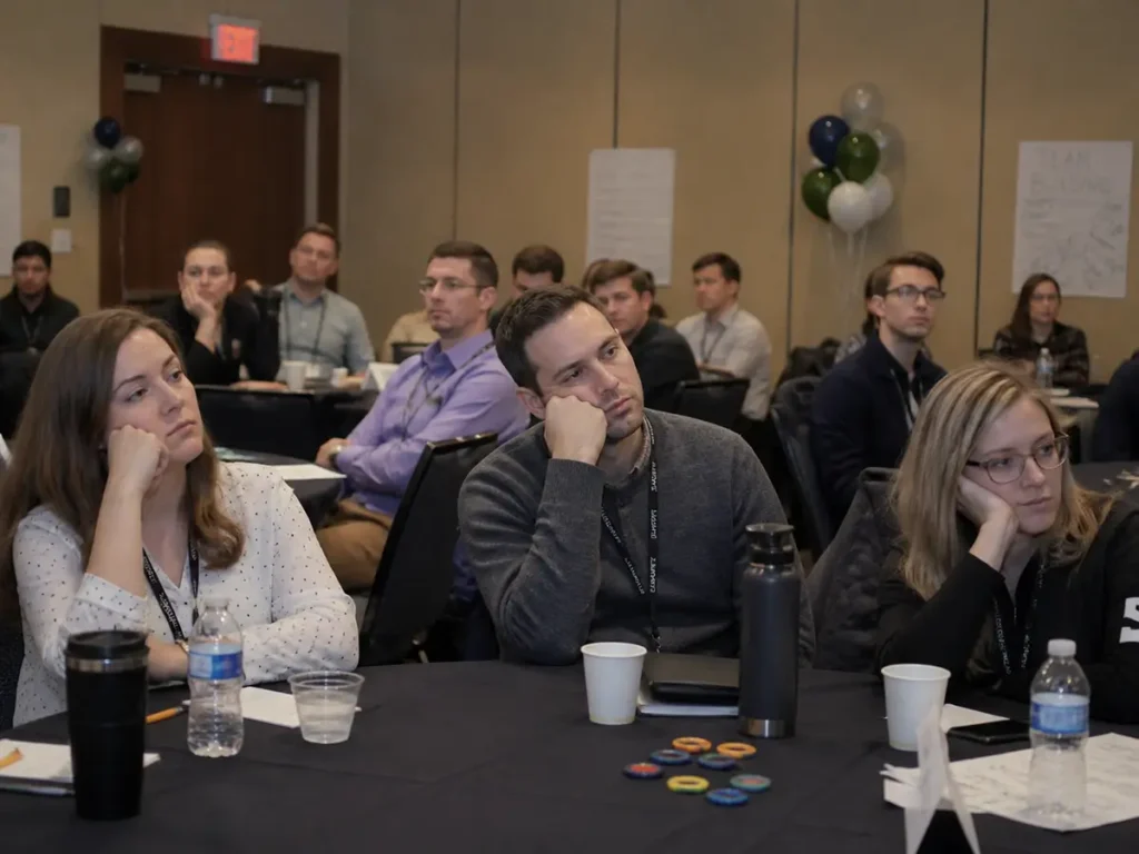 Conference attendees seated at tables listening attentively during a team meeting or workshop event, with balloons and flip charts in the background